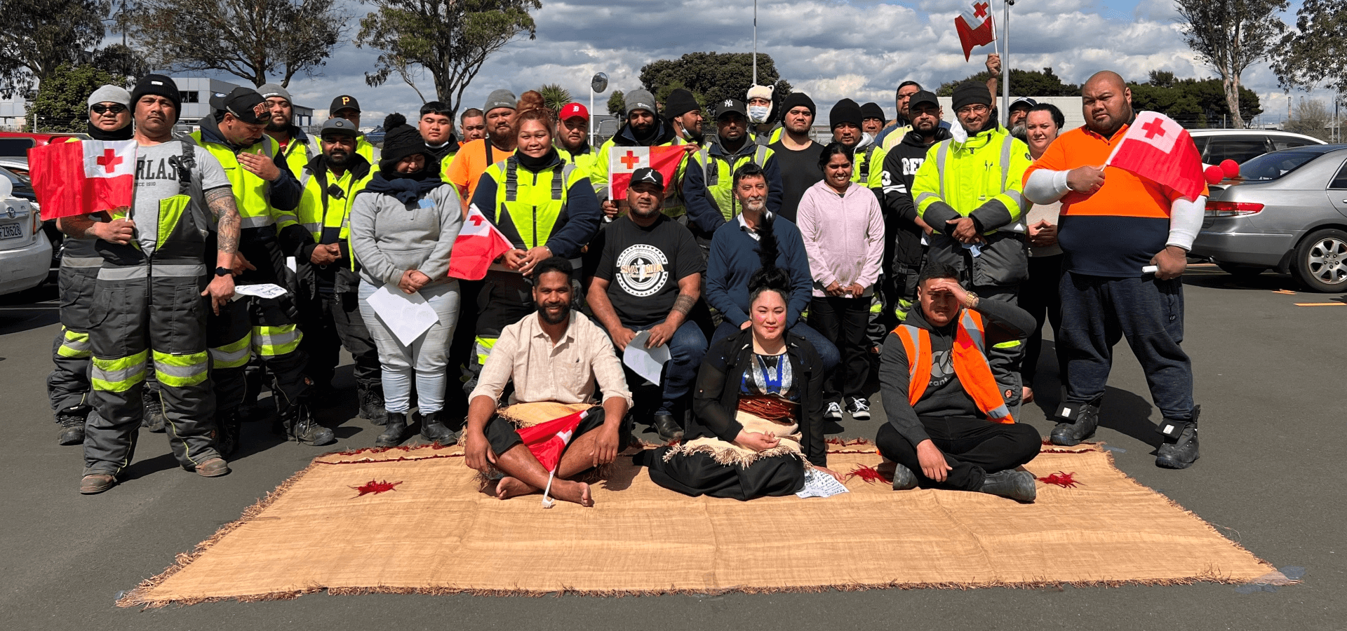 Foto de grupo de los miembros del equipo Lineage Auckland en Nueva Zelanda, reunidos al aire libre, mostrando la diversidad con varios miembros del equipo sosteniendo banderas y llevando chalecos de seguridad, reflejo de un lugar de trabajo multicultural.