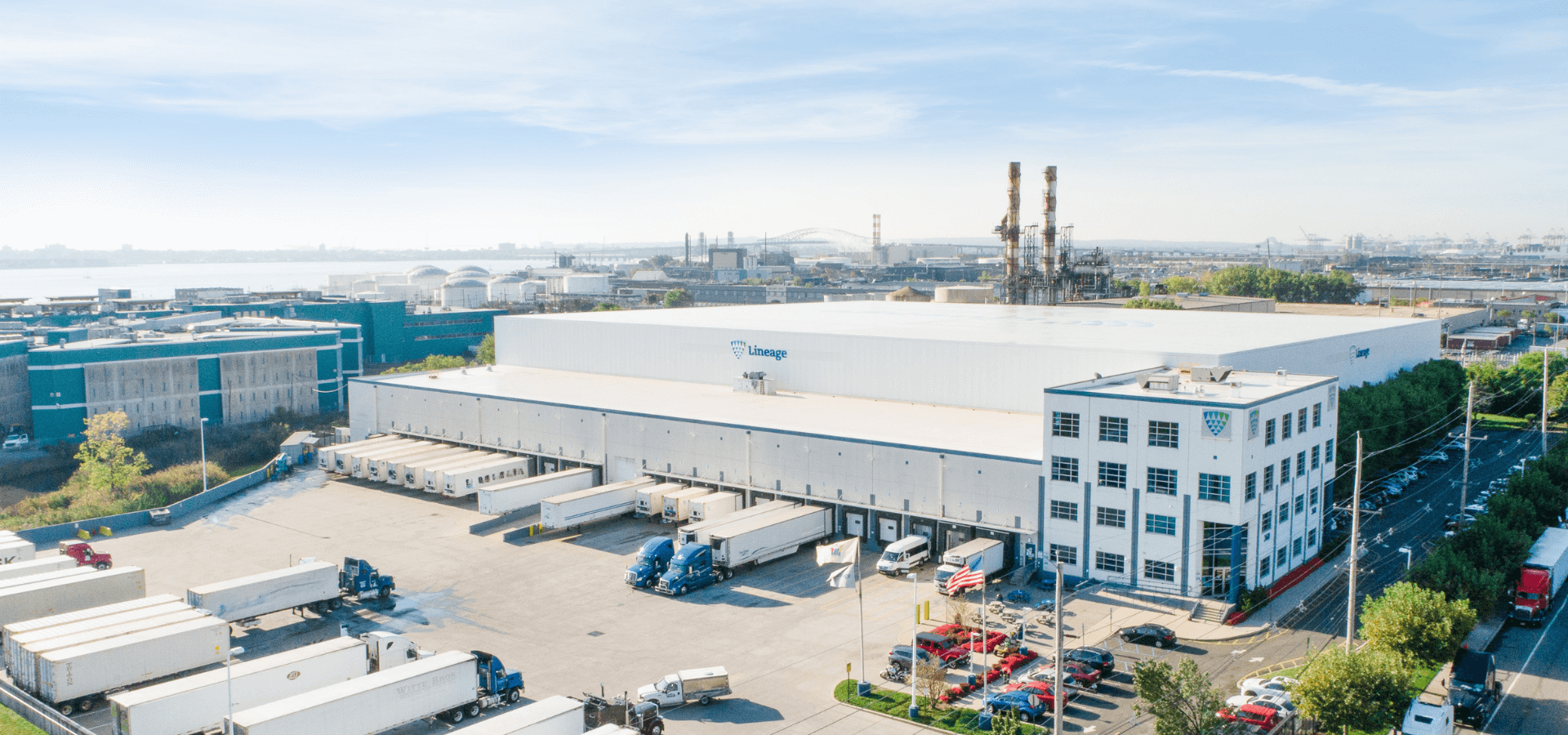 Un gran almacén frigorífico de Lineage en Nueva Jersey, con el logotipo de Lineage , con múltiples muelles de carga con camiones, situado en una zona industrial con proximidad a un puerto.