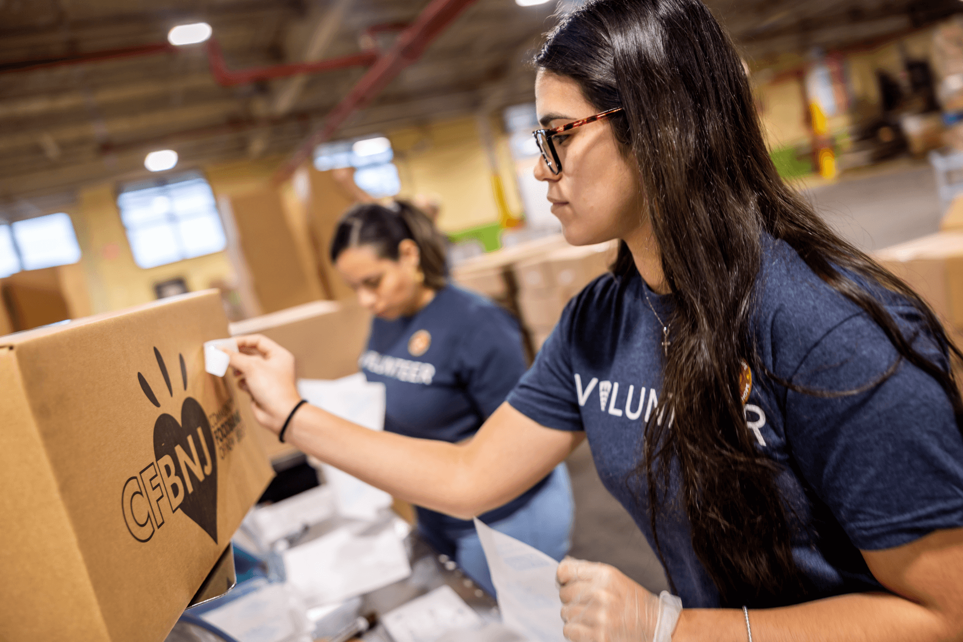 Dos voluntarios Lineage , con camisetas azules de "Voluntario", empaquetan y etiquetan cajas de alimentos en el interior de un almacén, preparando comida para donar a comunidades con inseguridad alimentaria.