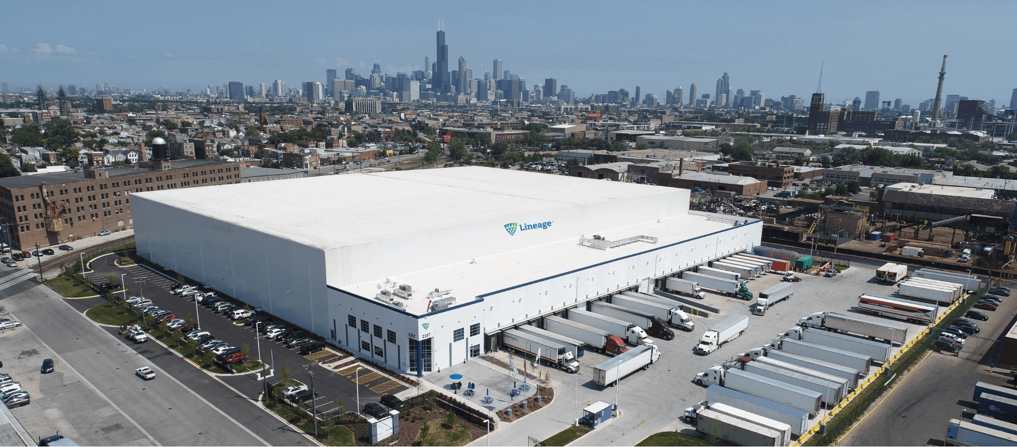 Vue aérienne de Lineageà Chicago, avec la ligne d’horizon de la ville en arrière-plan. De nombreux camions sont amarrés aux quais de chargement, ce qui met en valeur la capacité de l’installation pour la distribution à grande échelle. L’entrepôt est un bâtiment blanc avec le Lineage entouré d’aires de stationnement et de verdure.