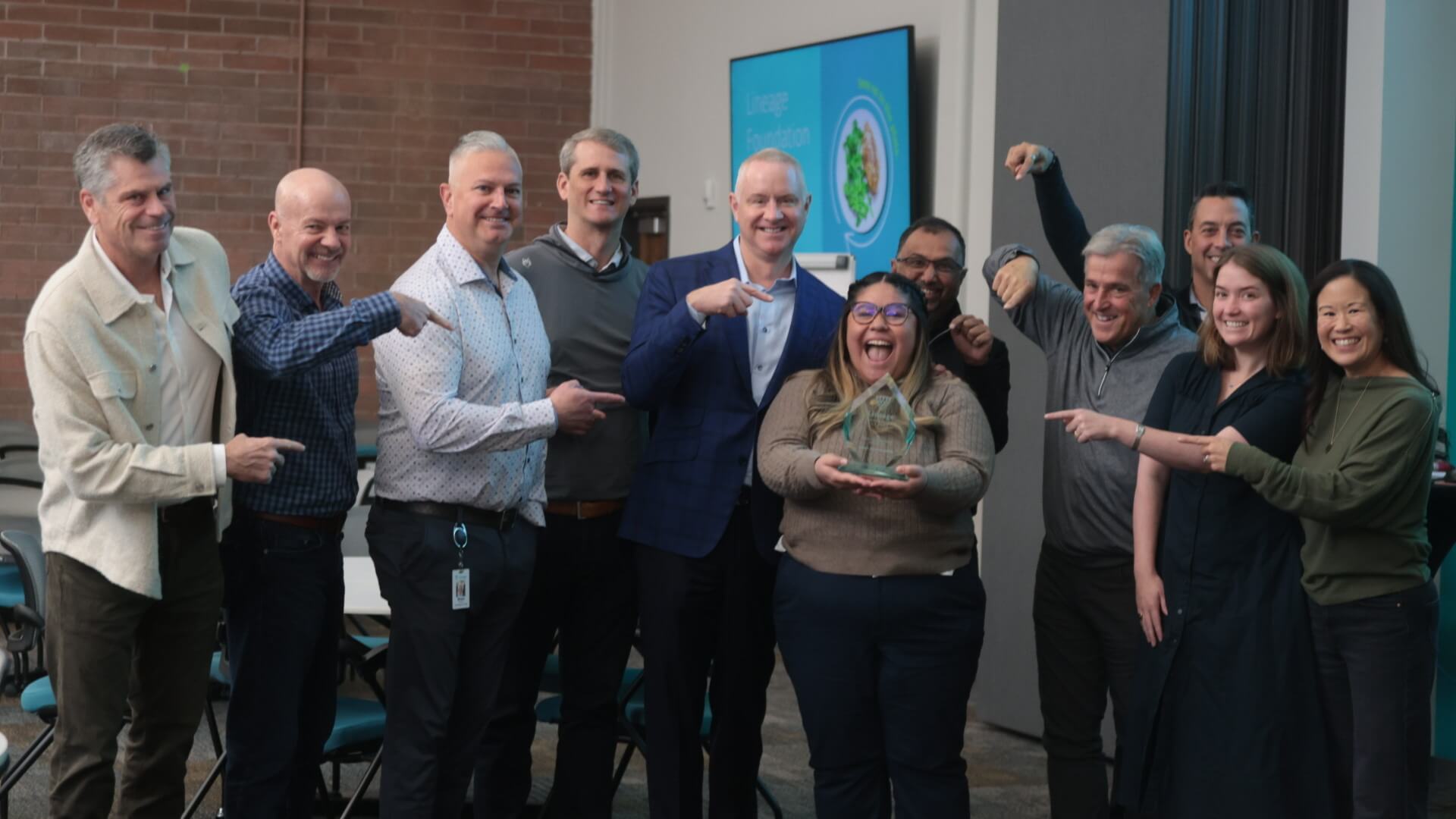 Swemy Sánchez Román, con su premio Servant Leader for Good en la mano, luce orgullosa mientras sus compañeros de Lineage celebran su logro, señalando y sonriendo en reconocimiento a su dedicación al servicio de la comunidad y al liderazgo.