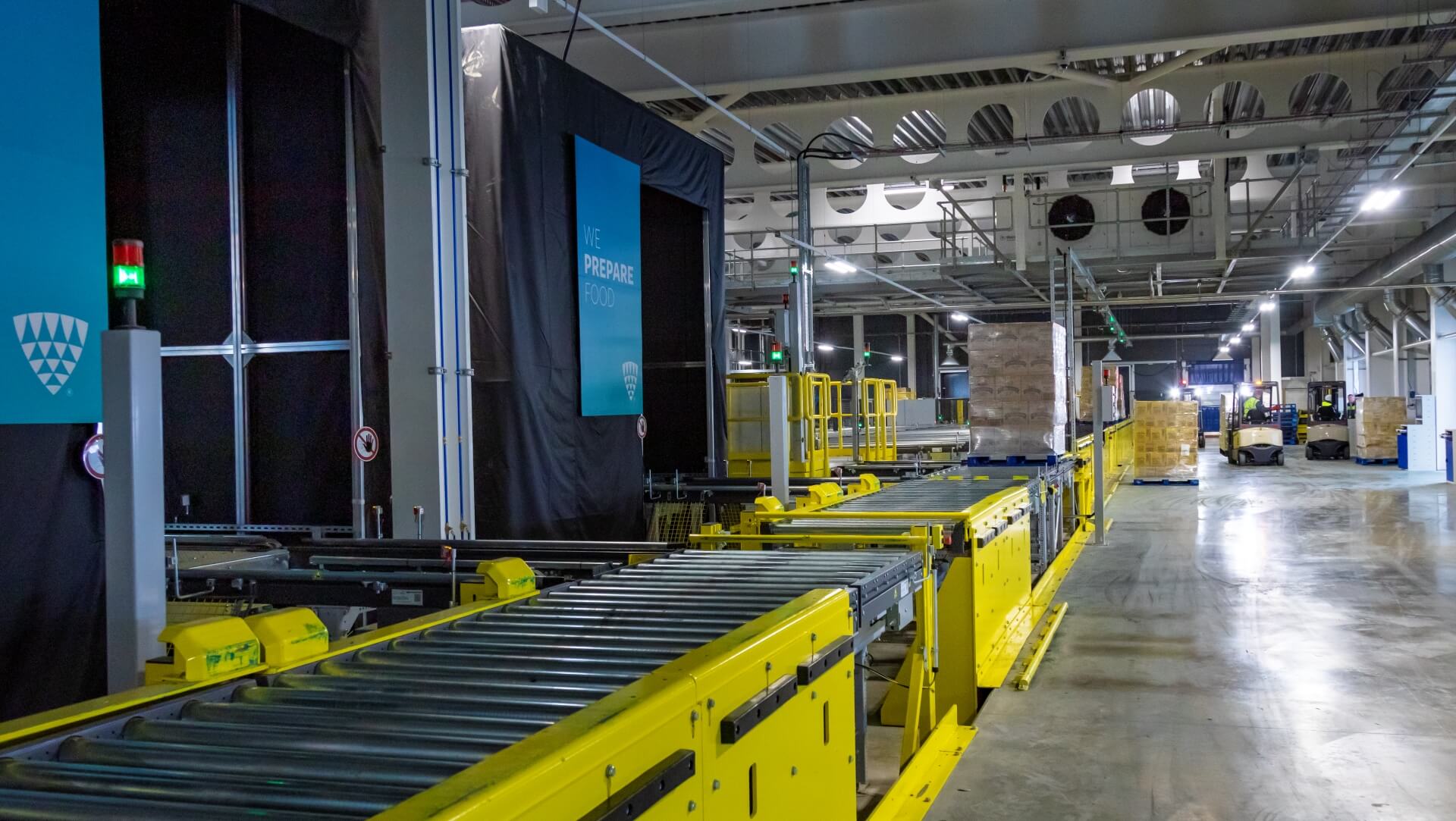 Un moderno almacén frigorífico con sistemas de transporte amarillos y carretillas elevadoras moviendo palés. La automatización Lineage Eye agiliza la recepción y la gestión del inventario.