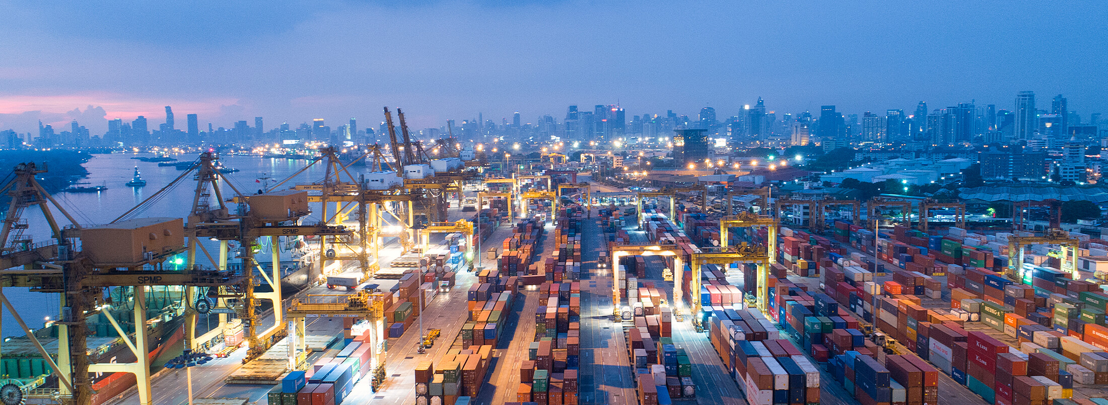 Aerial view of a busy port at dusk with shipping containers, cranes, and city skyline in the background, illustrating global trade and logistics operations.