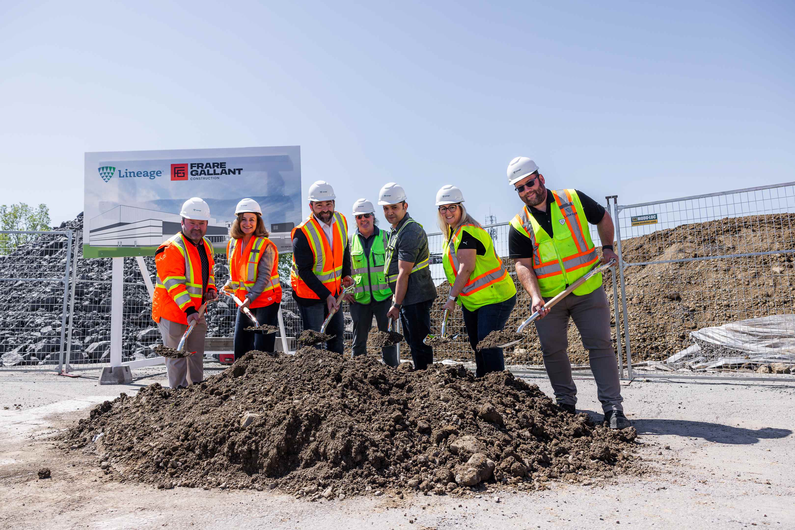 Un grupo de personas con cascos y chalecos de seguridad sonríen mientras sostienen palas en un montón de tierra en una obra.