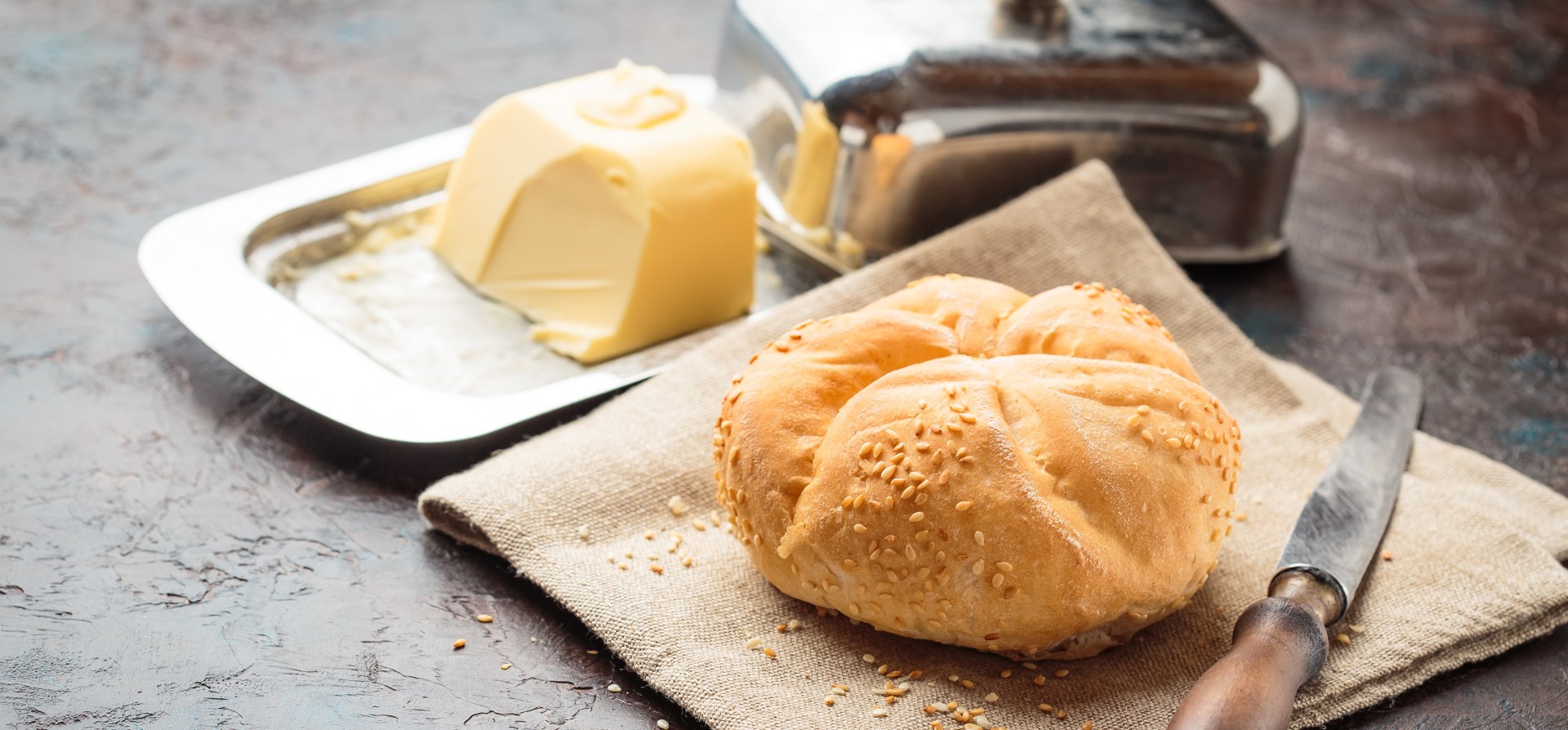 Fresh baked bread roll with sesame seeds on a cloth beside a butter dish and knife on a rustic kitchen surface