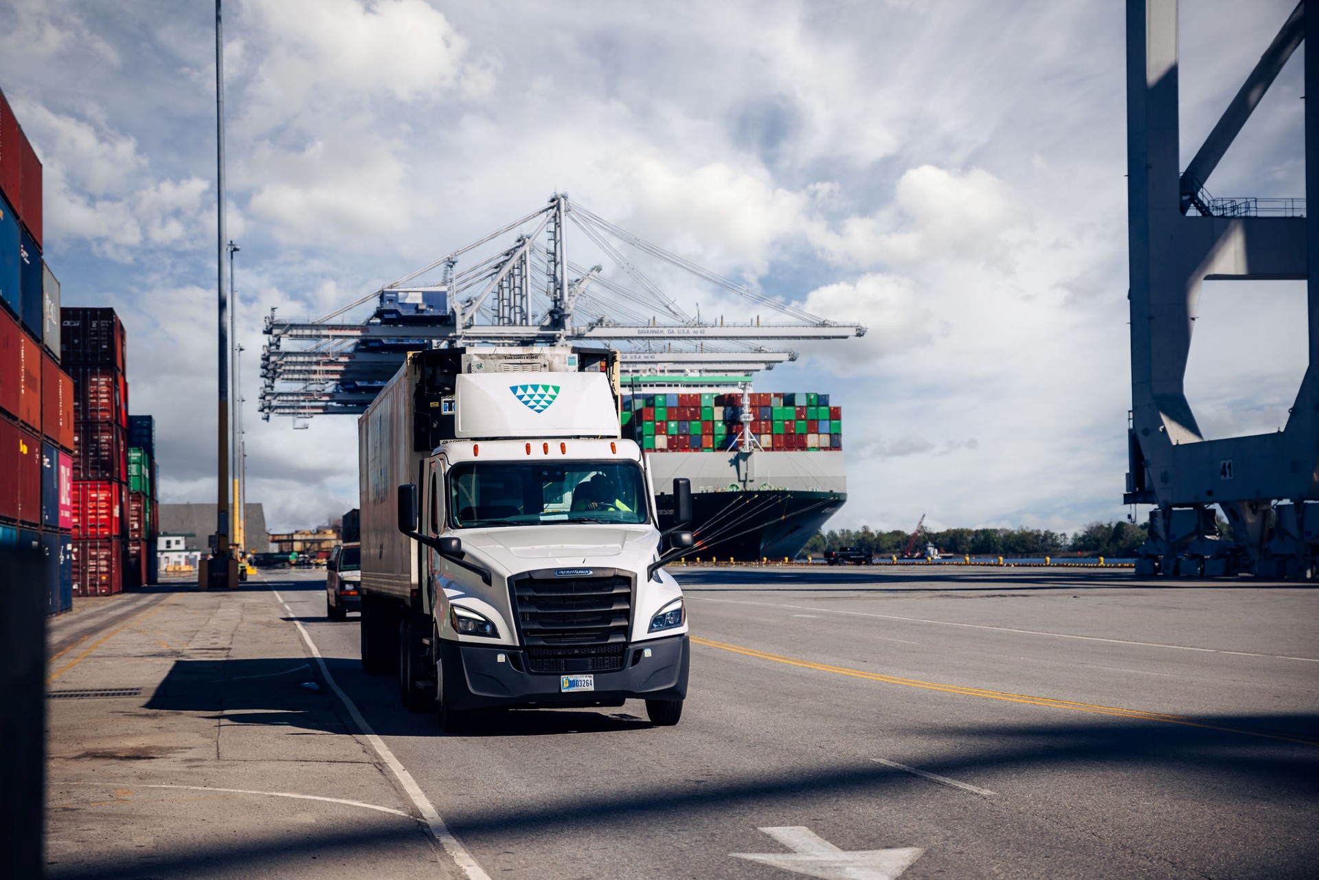 Lineage truck transporting refrigerated cargo through a container port with shipping cranes and cargo vessel in the background.