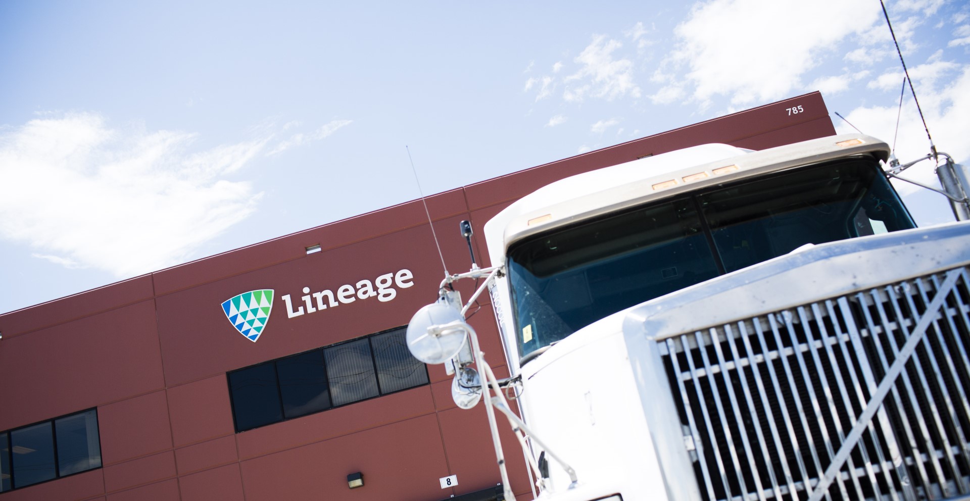 Lineage refrigerated truck parked outside a Lineage cold storage warehouse facility with loading dock doors and company signage visible.