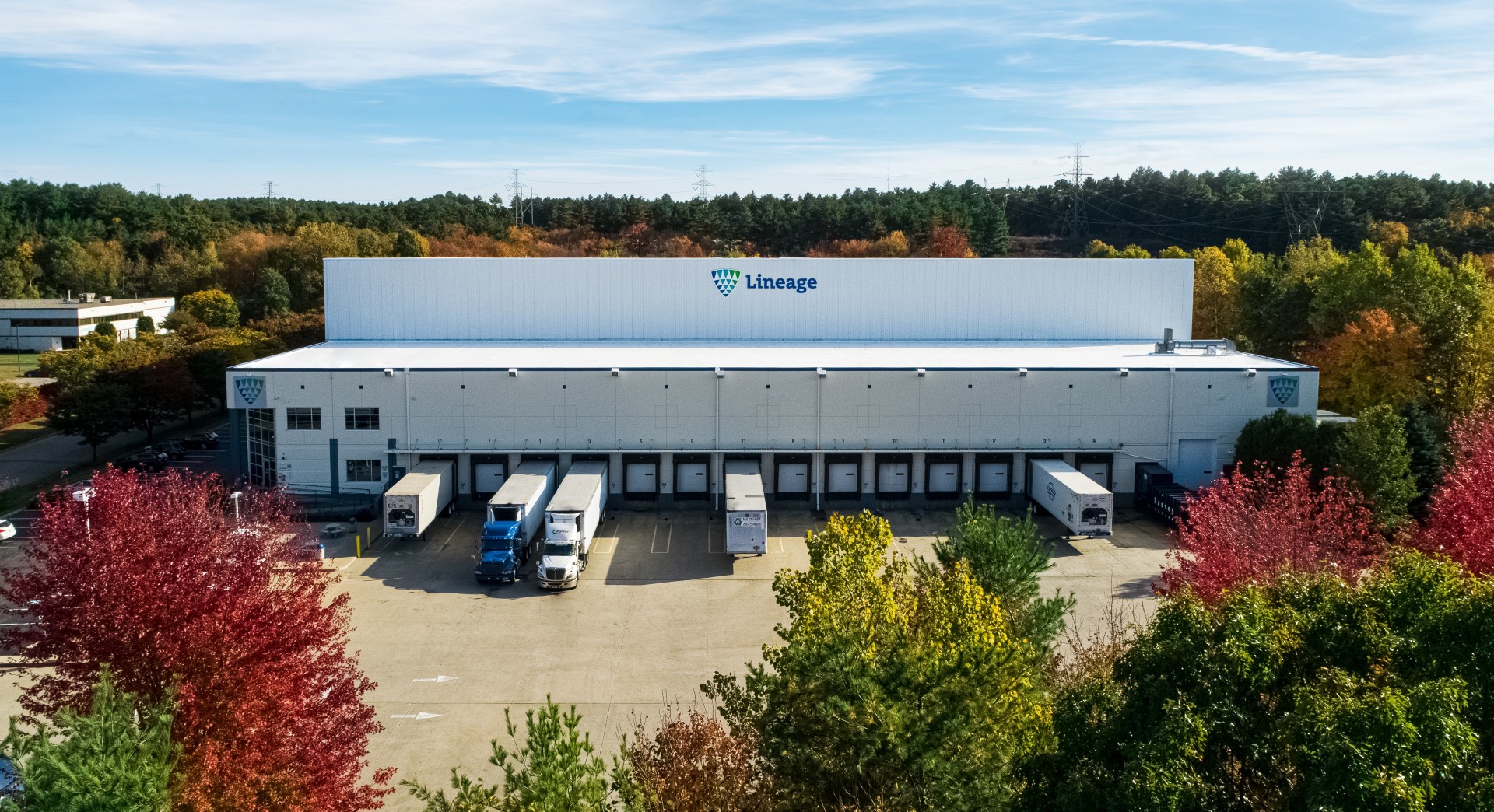 Aerial view of a Lineage cold storage facility in Massachusetts with trucks and trailers at loading docks, surrounded by autumn trees.