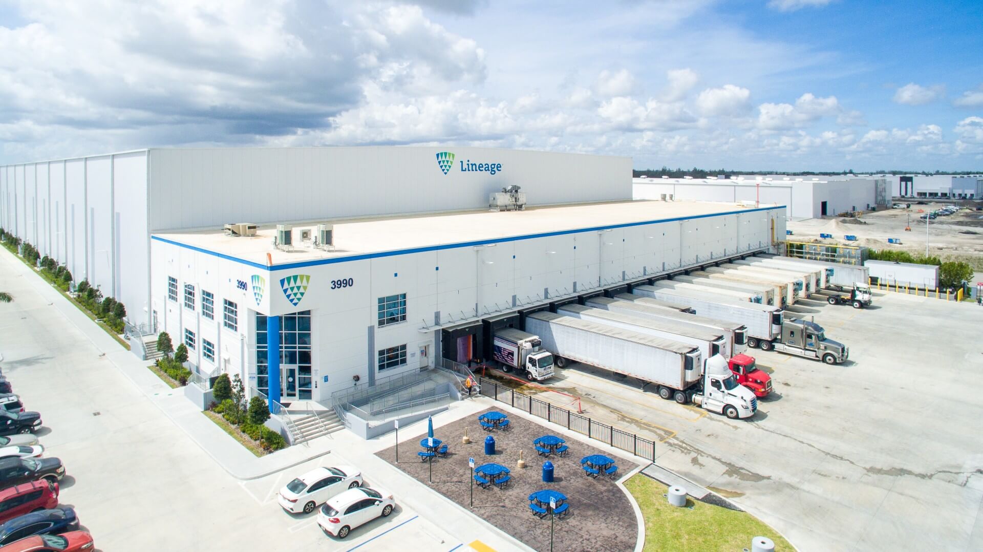 Aerial view of a Lineage cold storage warehouse with refrigerated trucks parked at loading docks and a parking area in the foreground.