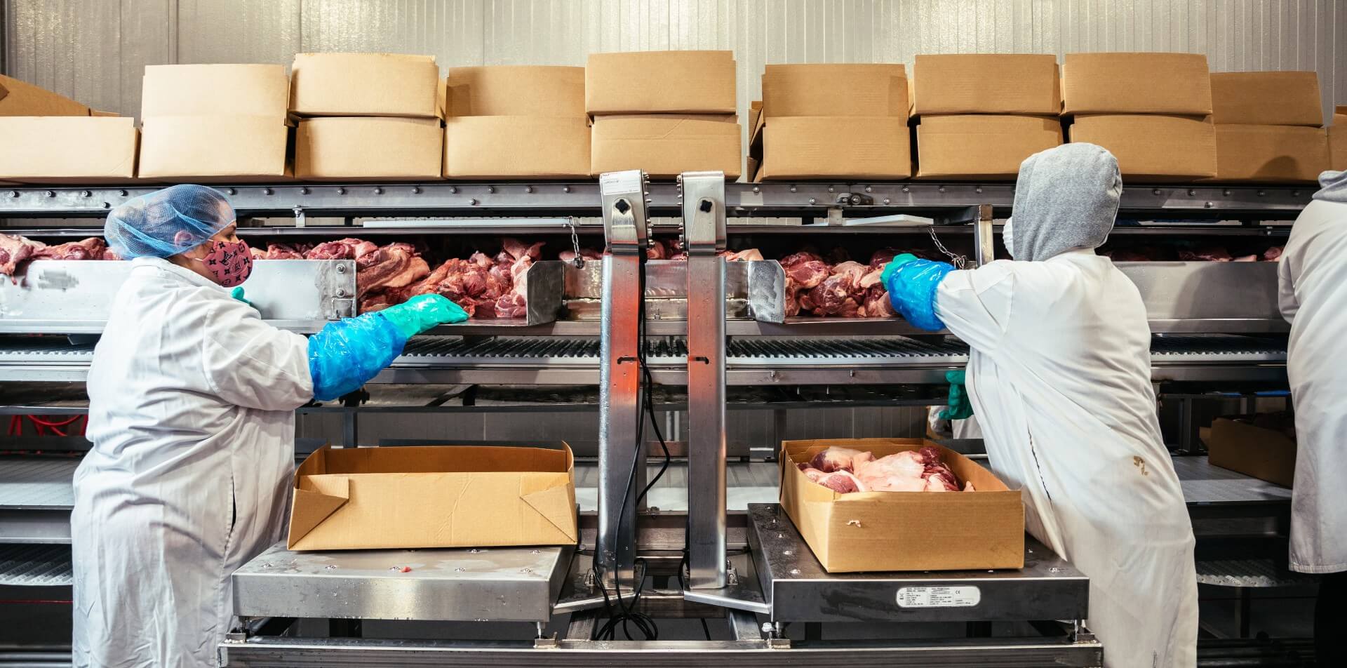 Two workers in protective clothing and gloves box meat products along a food processing line inside a Lineage facility.