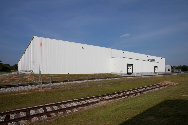 Vista lateral exterior del almacén frigorífico de Lineageen Macon, Atlanta, Georgia, con un almacén blanco, vías de tren adyacentes y un primer plano cubierto de hierba.