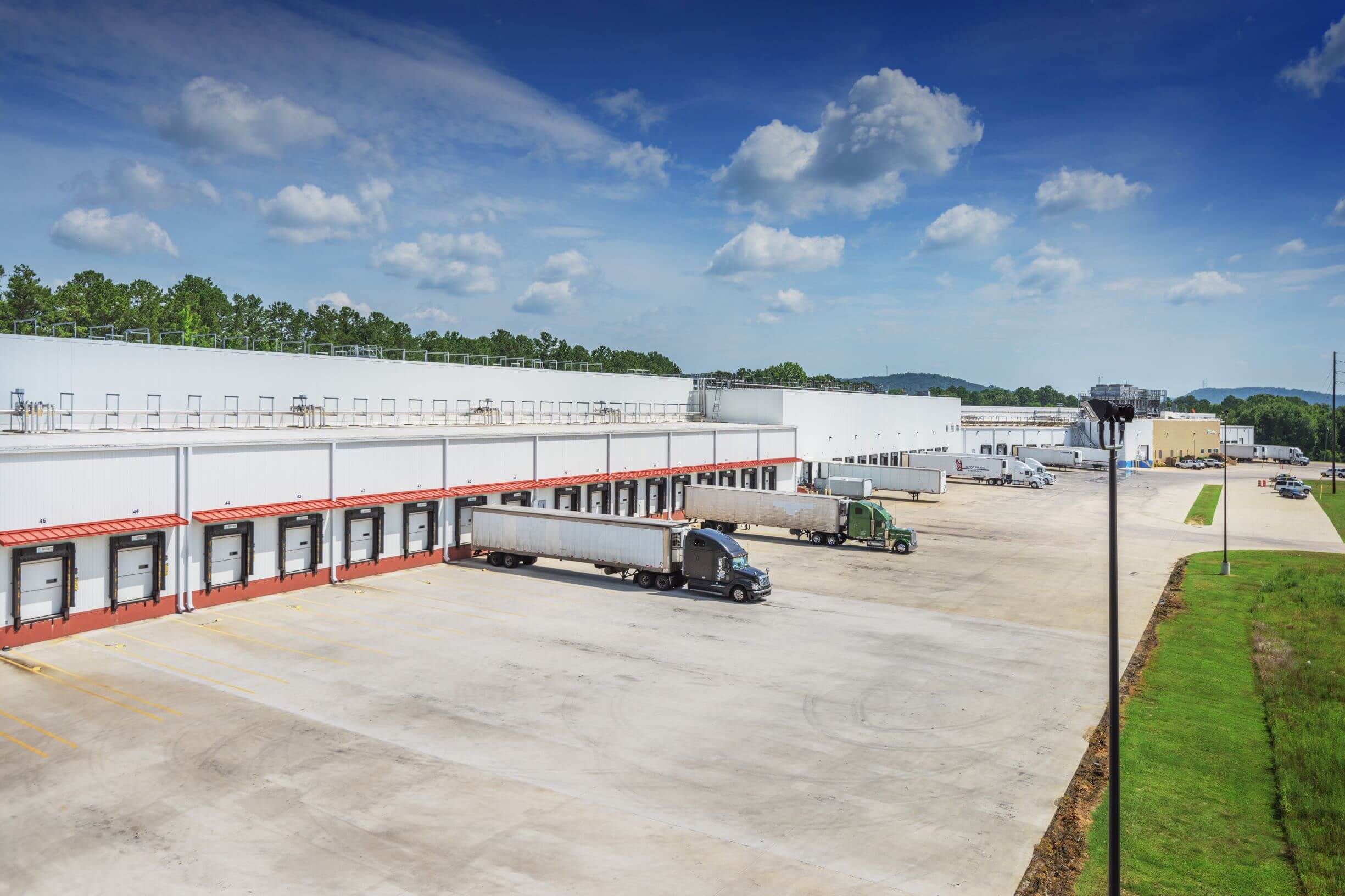 Vista del almacén frigorífico de Lineage en Attalla, Alabama, con varios muelles de carga y semirremolques aparcados para las entregas. La gran instalación blanca está rodeada por un extenso terreno de hormigón y un paisaje verde bajo un cielo azul brillante con nubes dispersas.
