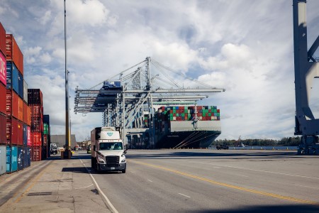 Lineage circulan por un puerto de contenedores junto a buques de carga y contenedores apilados, prestando apoyo a las operaciones de transporte de mercancías en régimen de depósito aduanero y a la logística de importación.
