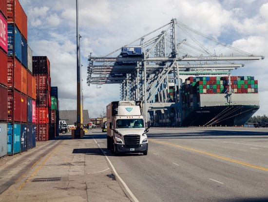 Lineage bonded drayage truck transporting refrigerated freight at a major shipping port with cargo containers, container cranes and an ocean cargo vessel in the background.