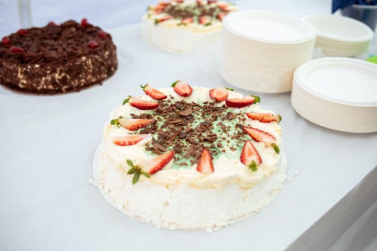 A decorated cream cake topped with sliced strawberries and chocolate pieces on a table beside plates.