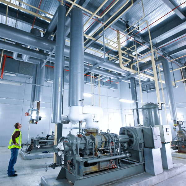 A worker in a reflective vest inspects the heavy-duty HVAC system and large piping inside a modern cold storage warehouse, with exposed ceiling conduits.