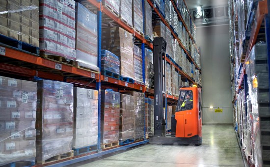 Forklift moving pallets of boxed goods in a temperature-controlled warehouse aisle with high storage racks