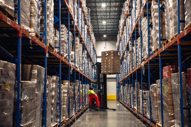 Forklift lifting a pallet of boxed products between tall storage racks inside a cold storage warehouse.