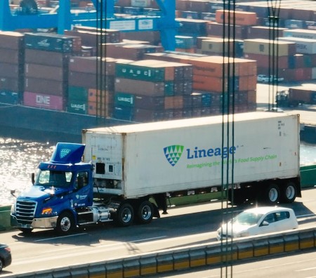 Lineage refrigerated truck traveling along a port-adjacent roadway with shipping containers visible in the background, supporting temperature-controlled freight movement.