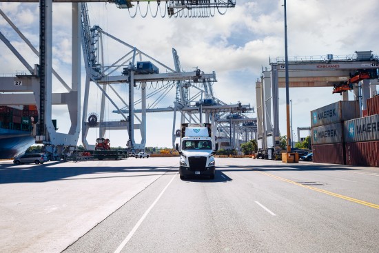 Refrigerated truck driving through a port terminal with cranes and shipping containers in the background