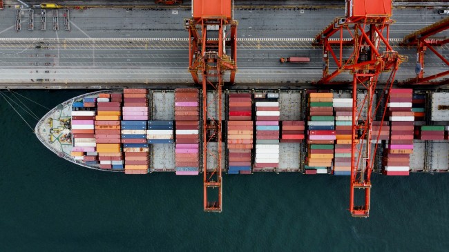 Overhead view of a cargo ship loaded with shipping containers beneath large port cranes at a shipping terminal.