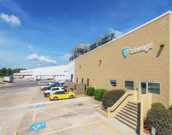 A Lineage cold storage warehouse in Attalla, Alabama, with the company's logo displayed on the beige exterior. The image shows a row of parked vehicles, including a yellow sports car, a loading dock area with semi-trucks, and the facility's infrastructure under a clear blue sky.