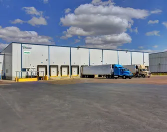 Exterior de las instalaciones de Lineageen McAllen West Military, mostrando el almacén blanco con múltiples muelles de carga y varios camiones aparcados bajo un cielo parcialmente nublado.