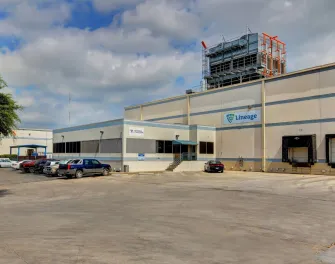 Vista exterior de las instalaciones de Lineageen San Antonio Profit, que muestra el almacén de color claro, los muelles de carga y los vehículos aparcados bajo un cielo parcialmente nublado.