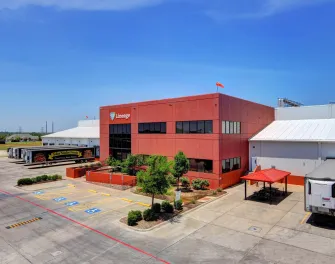 Vue extérieure d’un Lineage Installation de stockage frigorifique à Dallas, Texas, comprenant un immeuble de bureaux rouge, un entrepôt adjacent, des remorques réfrigérées stationnées et un ciel bleu clair.