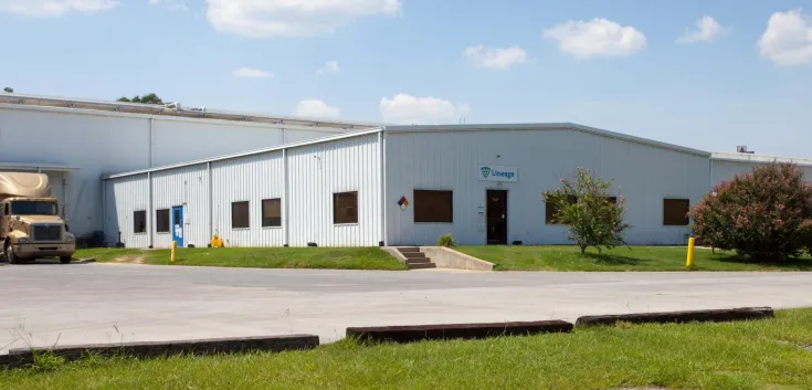 Vista exterior del almacén frigorífico de Lineageen Unadilla, cerca de Atlanta, Georgia, con un almacén blanco con una pequeña entrada de oficina, un camión aparcado y vegetación alrededor.