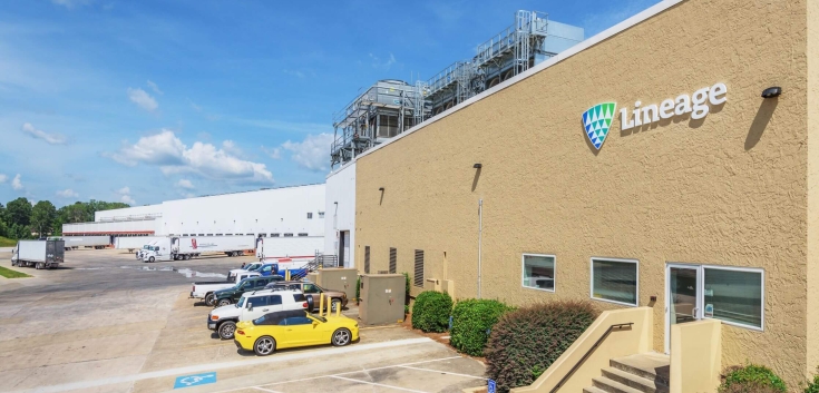 A Lineage cold storage warehouse in Attalla, Alabama, with the company's logo displayed on the beige exterior. The image shows a row of parked vehicles, including a yellow sports car, a loading dock area with semi-trucks, and the facility's infrastructure under a clear blue sky.