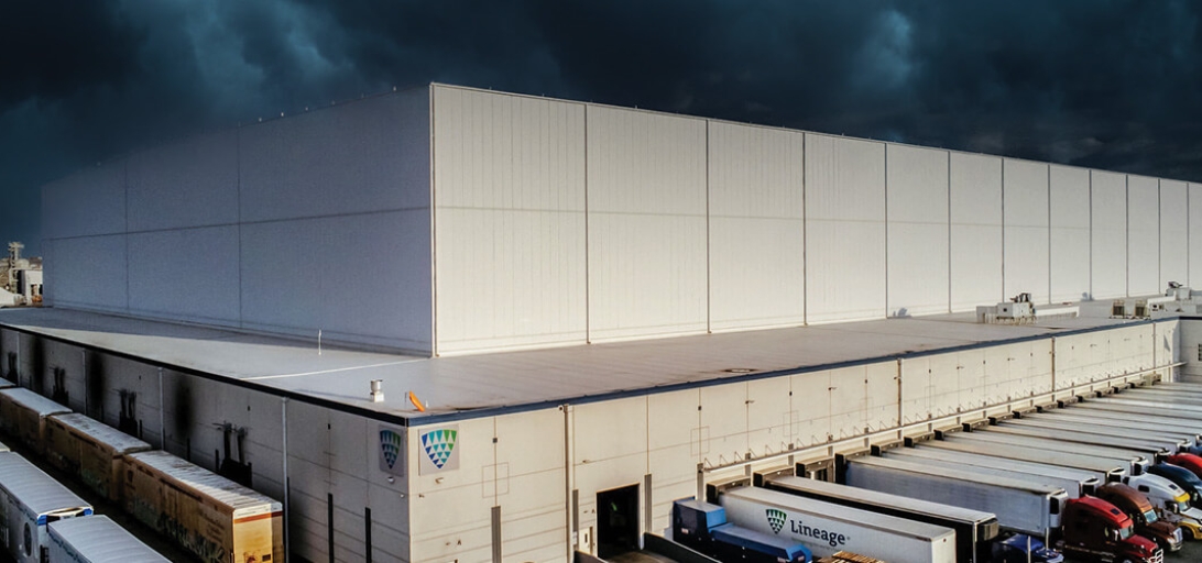 A Lineage warehouse under dark storm clouds, with multiple trucks and trailers docked, illustrating reliable cold chain logistics during severe weather conditions.