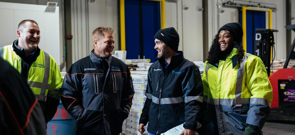 Group of team members laughing on warehouse floor during standup meeting