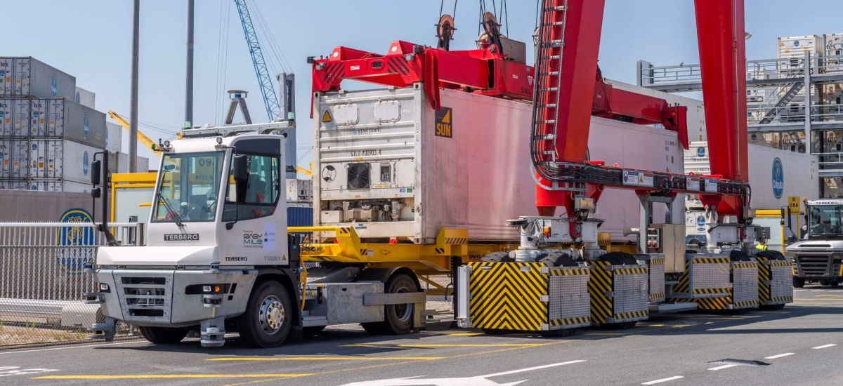 An autonomous terminal tractor by Terberg, equipped with sensor technology, is positioned under a towering red gantry crane handling a white shipping container, showcasing advanced logistics automation at a busy port.