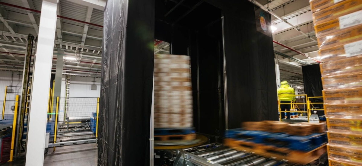A neatly stacked pallet positioned on a Lineage Eye conveyor system in a clean, well-organized, automated cold storage warehouse, with safety guardrails and scanning stations in the vicinity.