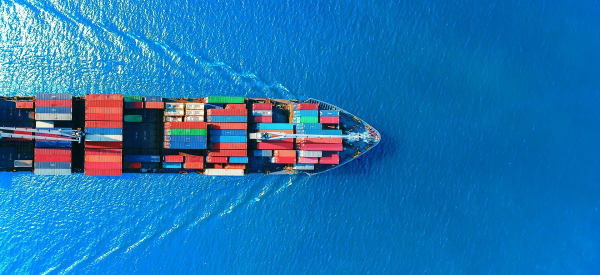 Aerial view of a large cargo ship carrying multicolored shipping containers across a bright blue ocean, symbolizing global trade and port logistics.