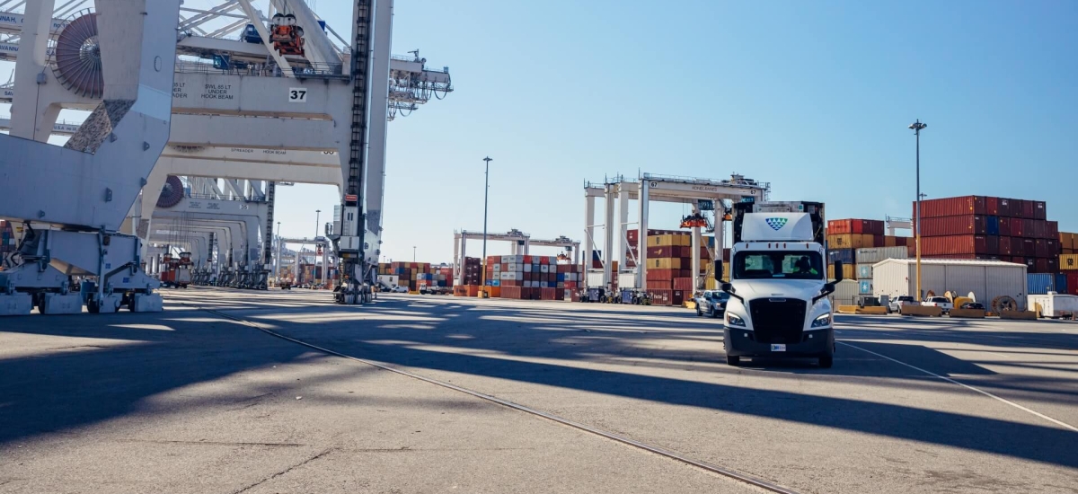 Lineage truck driving through a container port surrounded by cranes and stacked shipping containers, highlighting import/export and drayage operations
