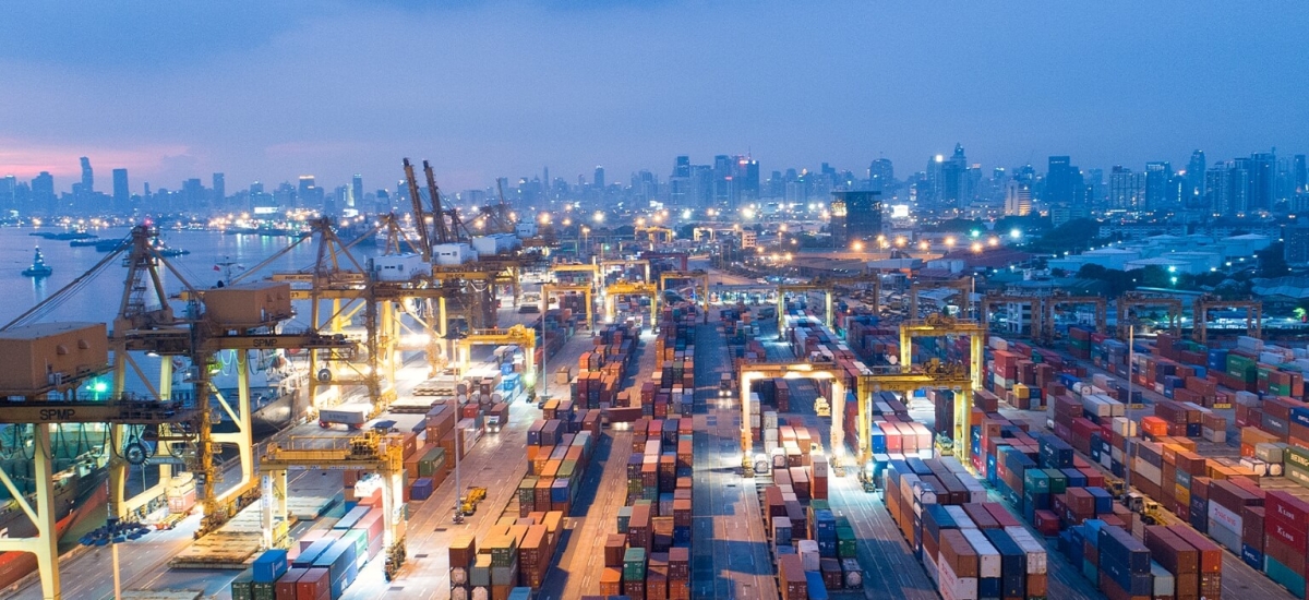 Aerial view of a busy shipping port at dusk, with rows of colorful cargo containers, large container cranes and a lit-up city skyline in the background.