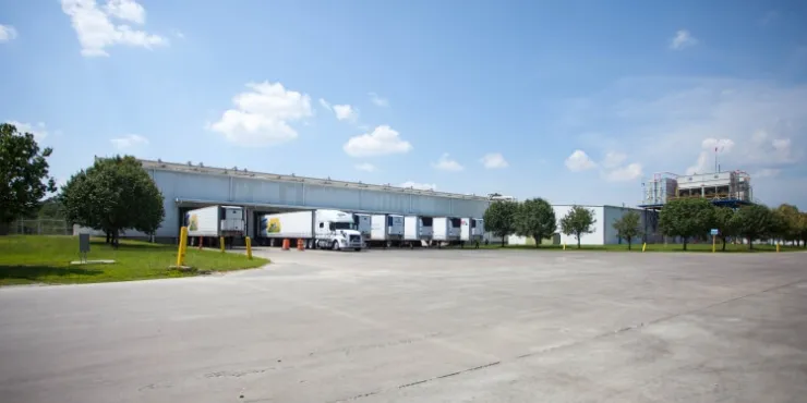 Vista lateral del almacén frigorífico de Lineageen Unadilla, cerca de Atlanta (Georgia), con los remolques frigoríficos atracados, la vegetación circundante y zonas de aparcamiento abiertas bajo un cielo azul despejado.