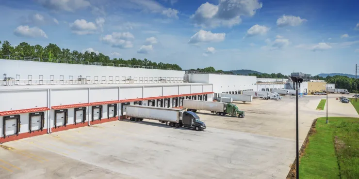 Vista del almacén frigorífico de Lineage en Attalla, Alabama, con varios muelles de carga y semirremolques aparcados para las entregas. La gran instalación blanca está rodeada por un extenso terreno de hormigón y un paisaje verde bajo un cielo azul brillante con nubes dispersas.