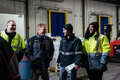 Team members gathered, laughing at standup meeting on warehouse floor.