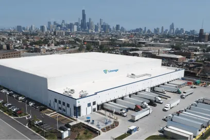 Aerial view of Lineage’s cold storage warehouse in Chicago, with the city skyline in the background. Numerous trucks are docked at loading bays, showcasing the facility’s capacity for large-scale distribution. The warehouse is a white building with the Lineage logo, surrounded by parking areas and greenery.
