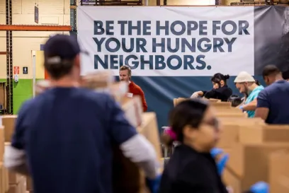 Volunteers packing boxes of food in a warehouse, with a large banner reading "Be the hope for your hungry neighbors" in the background.