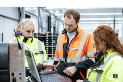 Tres miembros del equipo Lineage con chalecos de seguridad de alta visibilidad colaboran en un almacén, revisando datos en un ordenador para coordinar la logística de la cadena de frío.
