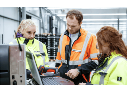 Three Lineage team members in high-visibility safety jackets collaborate in a warehouse, reviewing data on a computer to coordinate cold chain logistics.