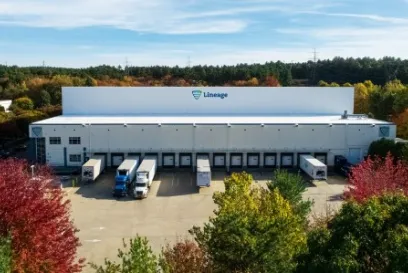 Aerial view of a Lineage cold storage facility in Massachusetts with trucks and trailers at loading docks, surrounded by autumn trees.