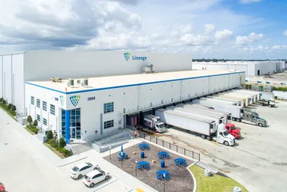 Aerial view of a Lineage cold storage warehouse with refrigerated trucks parked at loading docks and a parking area in the foreground.