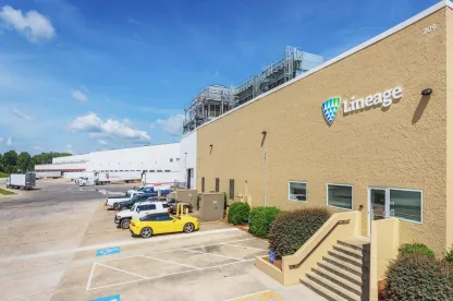 Un almacén frigorífico de Lineage en Attalla, Alabama, con el logotipo de la empresa en el exterior beige. La imagen muestra una fila de vehículos aparcados, entre ellos un deportivo amarillo, una zona de muelles de carga con semirremolques y la infraestructura de las instalaciones bajo un cielo azul despejado.